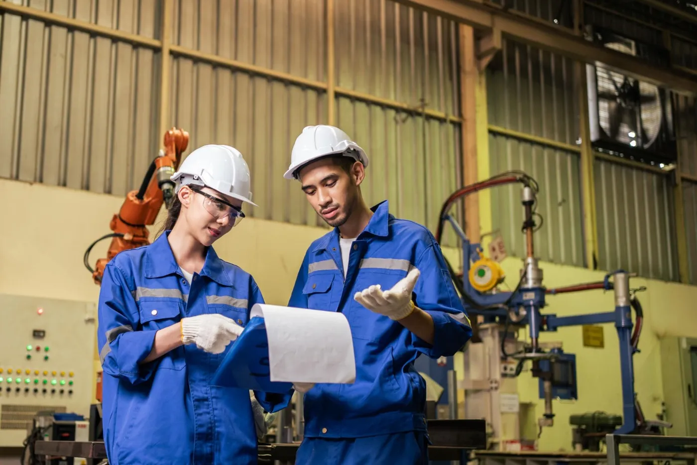 Two manufacturing workers in hard hats reviewing production documents beside a robotic arm