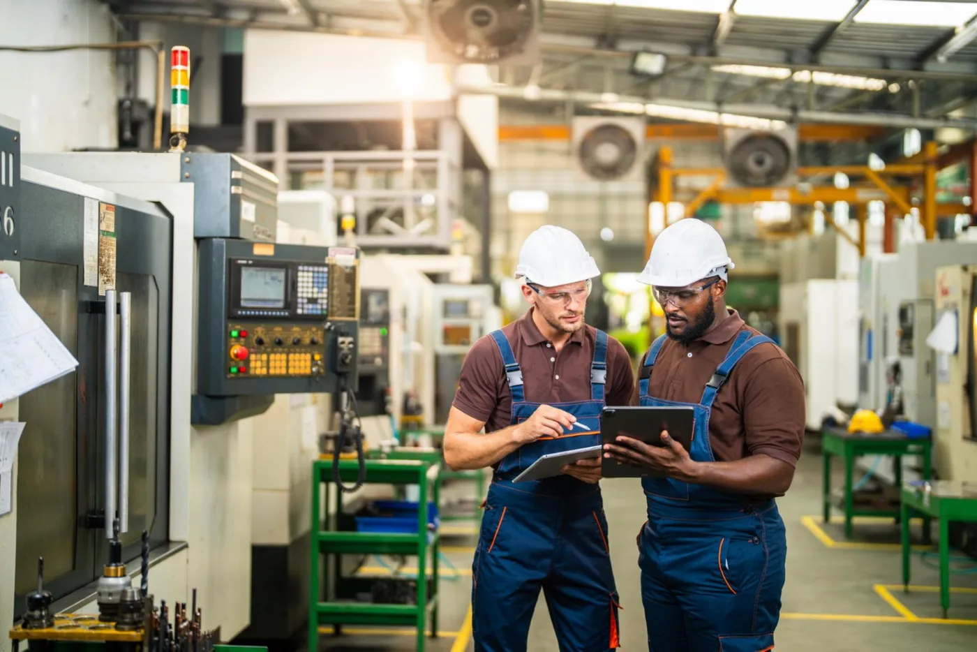 Two technicians with tablets comparing notes on an industrial plant floor beside CNC machinery
